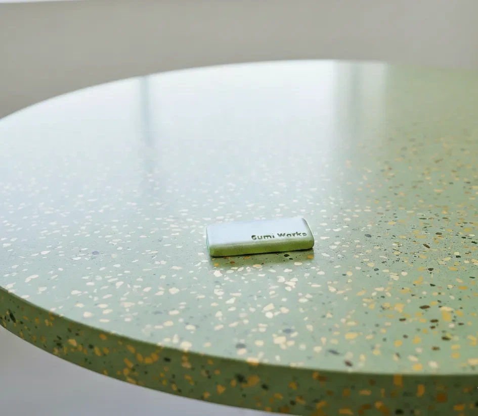 Round dining table close-up view. A statement table made from forest-green colour terrazzo concrete, using natural golden colour hues stone aggregates. Gumi Works' London studio colour sample on table, as a reference to concrete colour matching in production.